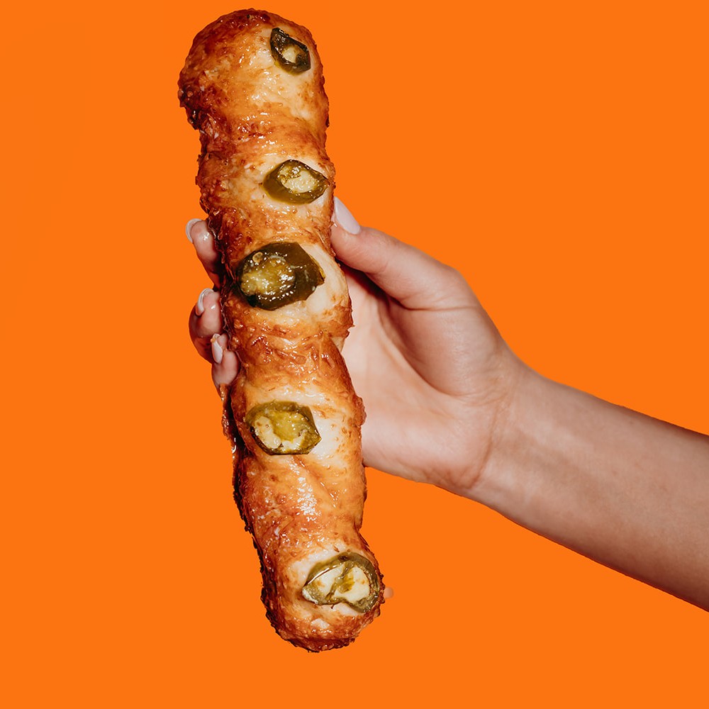 A hand holding a Supreme Pretzel Twist topped with jalapeno slices and melted Monterey Jack cheese