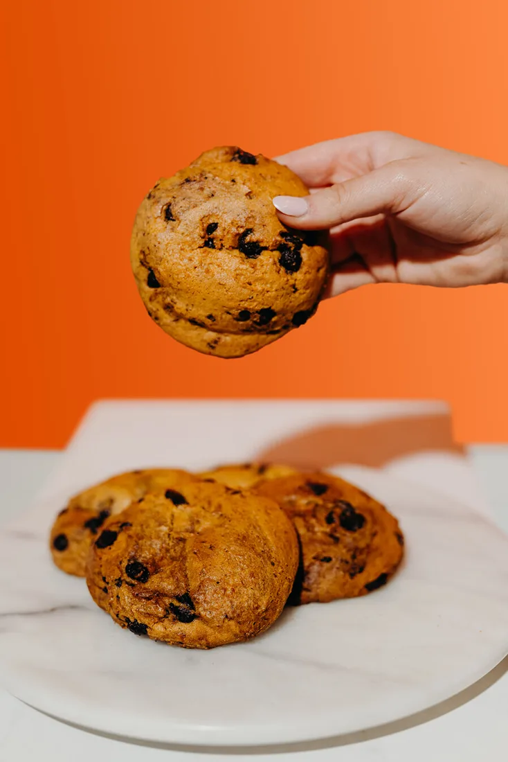 A hand holding a thick Thirst pumpkin chocolate chip cookie above a plate of more cookies