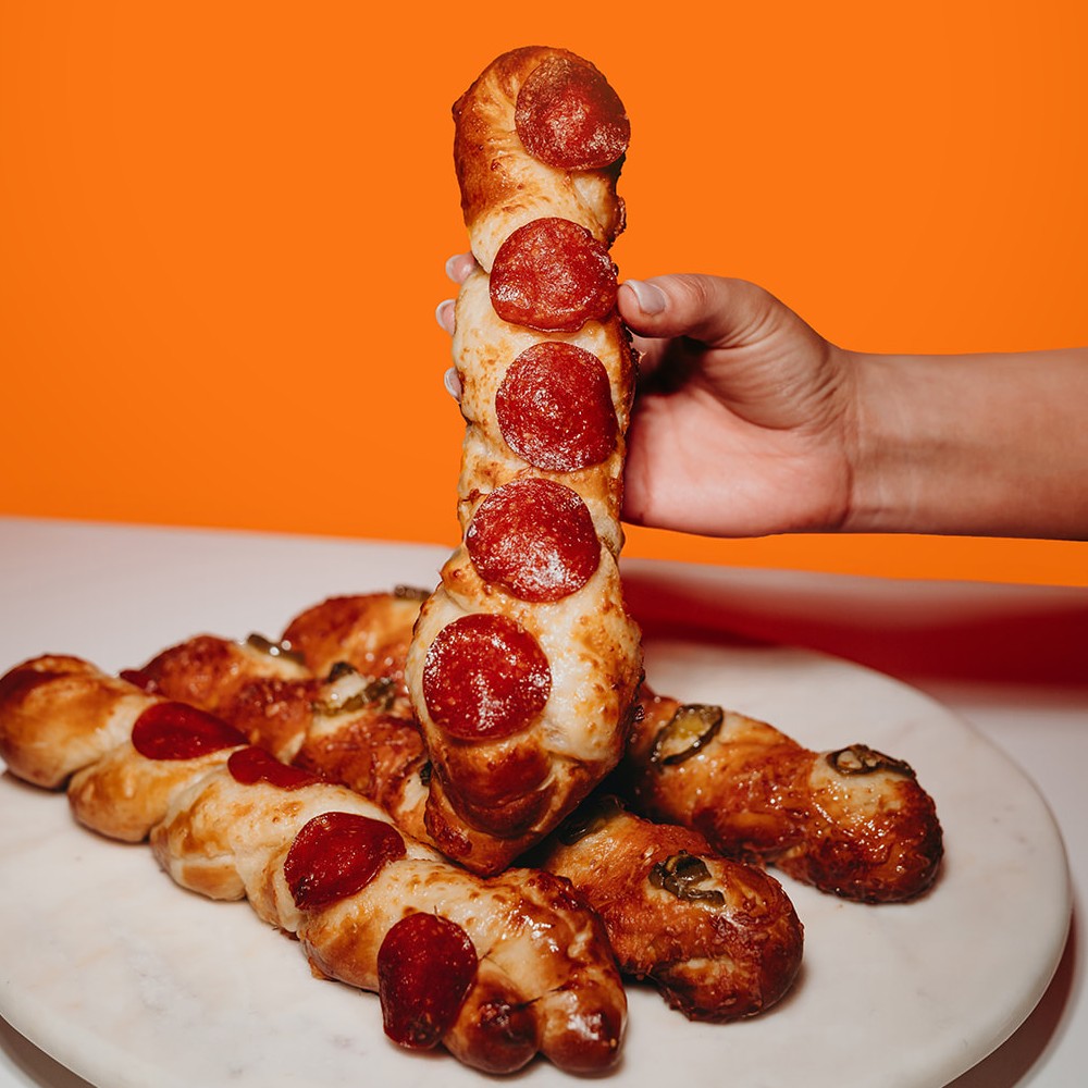 A hand holding a pepperoni Supreme Pretzel Twist above a marble platter piled with pretzel twists